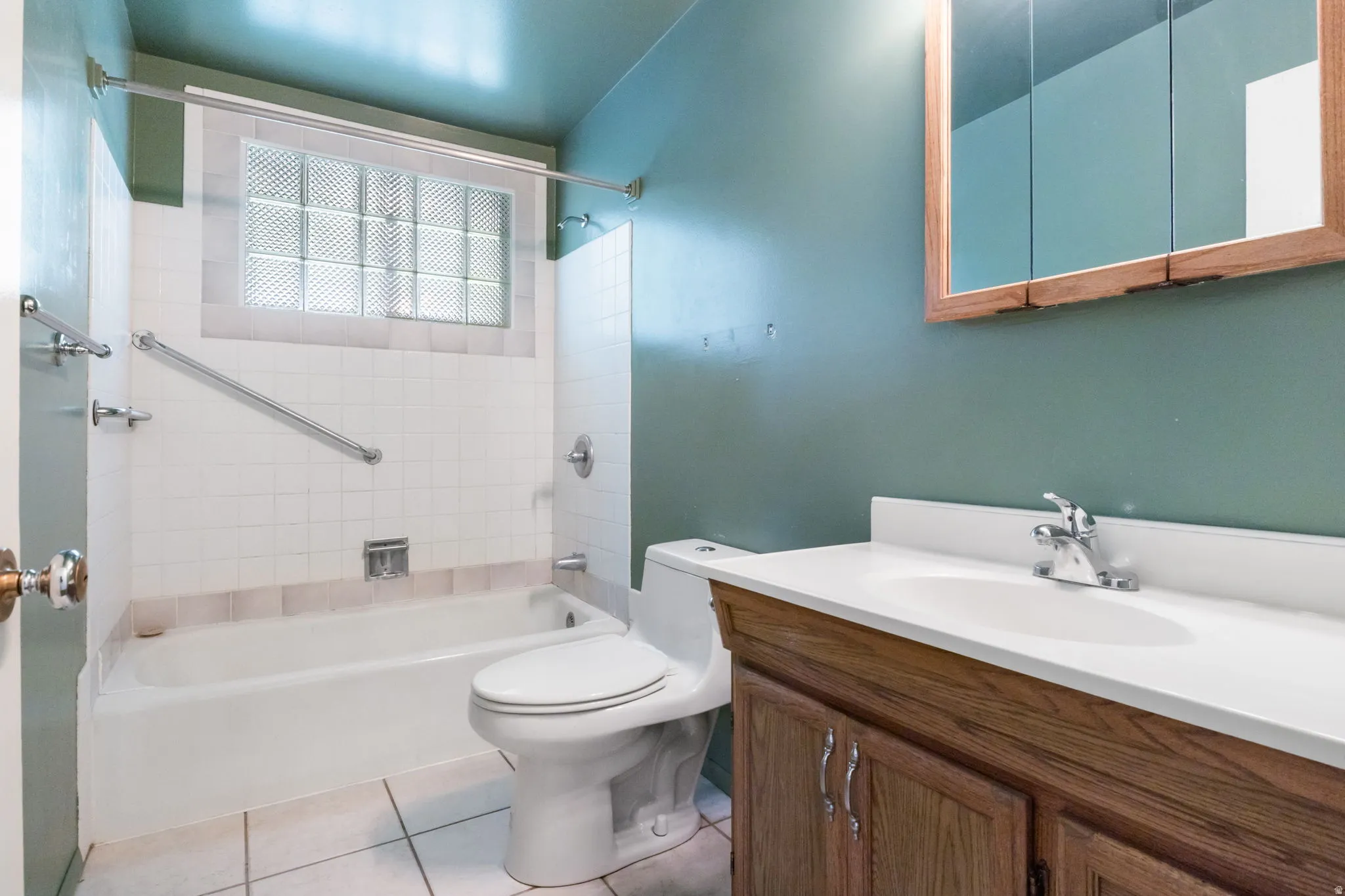 Bathroom featuring a built-in tub, white tile surround, glass block window, wood-finish vanity with an integrated sink, and a mirrored medicine cabinet