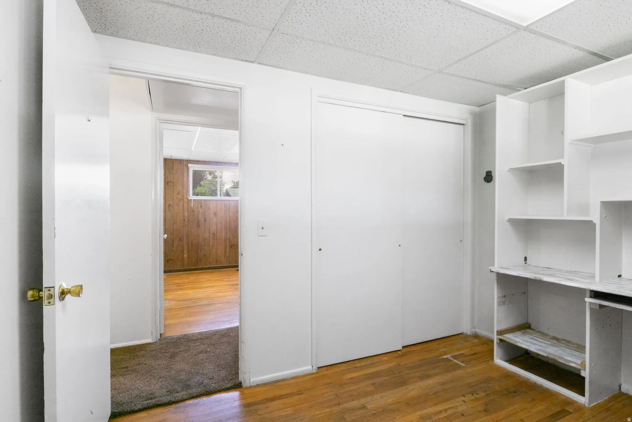 Hardwood-style flooring, white built-in shelving, white sliding closet doors, white walls, and a dropped ceiling