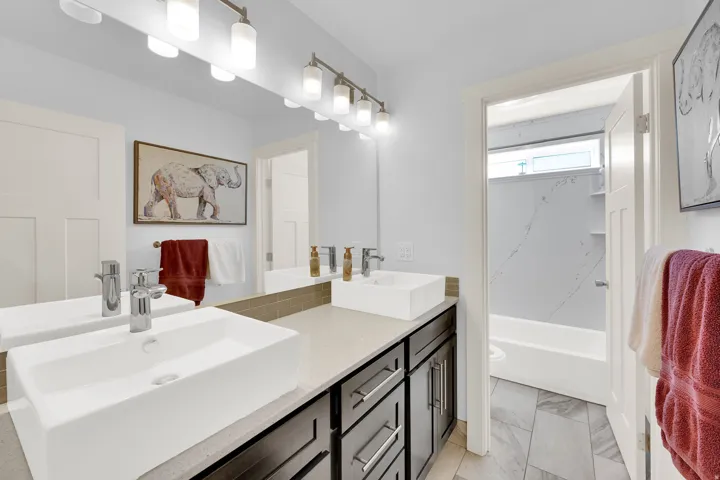 Dual vessel sinks with chrome fixtures, dark wood-finish vanity with storage, stone-look countertop, full-width mirror, and gray wood-look flooring