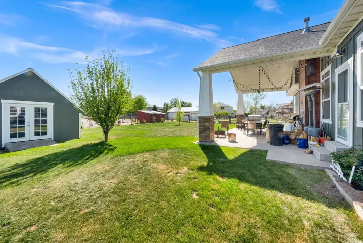 Expansive lawn with a mature tree, covered patio featuring stone-accented pillars, and a detached outbuilding with French doors