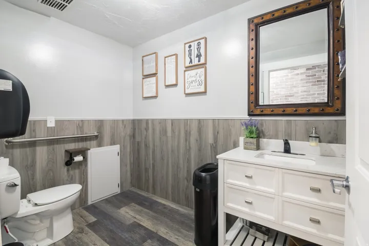 Wood-finish flooring, wood-look wainscoting, white walls, white vanity with a white countertop and integrated sink, and a dark-framed mirror