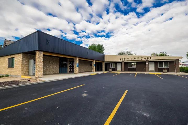 Commercial building with a brick facade, vertical panel siding, covered walkway, multiple storefront entrances, and an asphalt parking lot with painted yellow stripes
