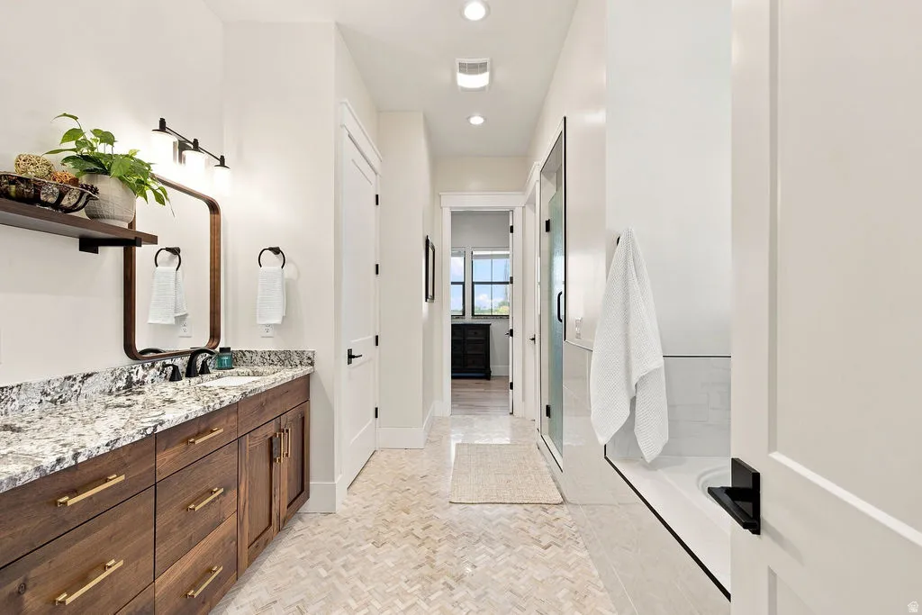 Spacious bathroom featuring a double vanity with granite countertop, dark wood-finish cabinetry with gold-tone hardware, a walk-in shower with clear glass enclosure, and light-toned mosaic floor tiling