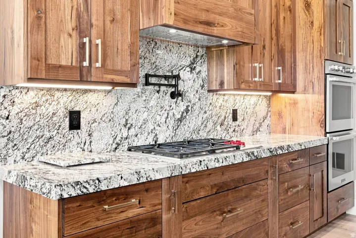 Wood-finish cabinetry with polished hardware, stone countertops with matching backsplash, built-in gas cooktop, and double wall ovens