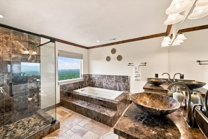 Dual vessel sinks with bronze-finish fixtures, a large soaking tub with natural stone surround, and a glass-enclosed shower with natural stone tile