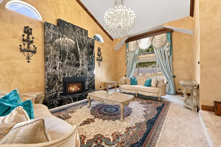 Vaulted ceiling with an ornate crystal chandelier, dark marble-clad fireplace, arched transom windows, and custom drapery