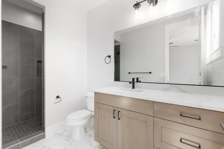 Walk-in shower with dark gray tile, light wood-finish vanity with dual sinks, white countertop, matte black hardware, and large vanity mirror