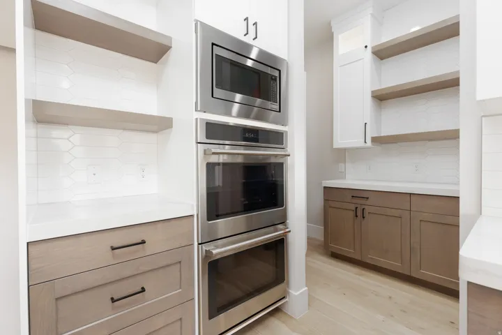 Stainless steel built-in microwave and double ovens, light wood-finish flooring, white countertops, two-tone cabinetry, and an elongated hexagon tile backsplash