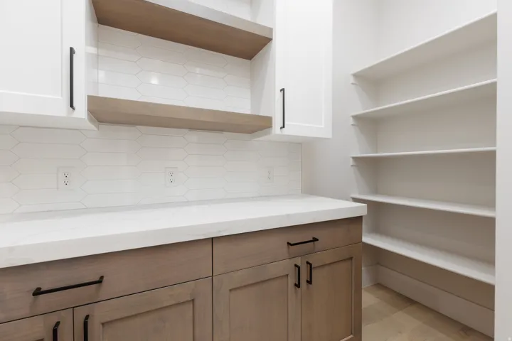 Built-in pantry shelving, lower wood-finish cabinetry, upper white cabinetry, light-colored countertops, and a white geometric tile backsplash