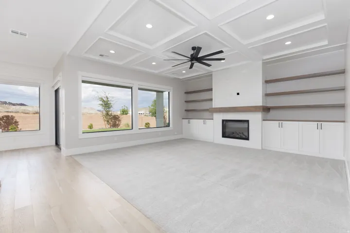Coffered ceiling with recessed lighting and a ceiling fan, a built-in fireplace with a wood mantle, custom cabinetry, integrated shelving, and large picture windows