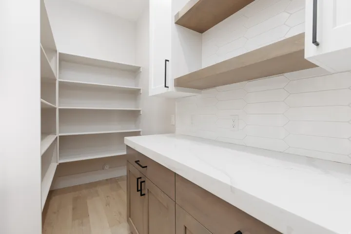 Custom built-in pantry shelving, spacious white countertop, wood-finish flooring, contemporary wood-finish cabinetry, and a geometric tile backsplash