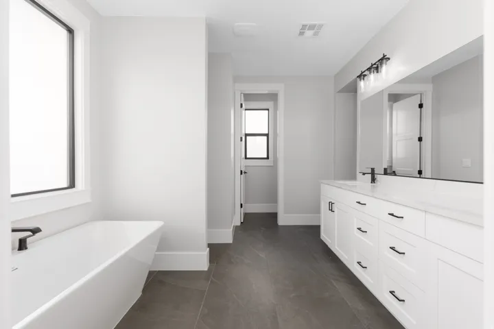 Freestanding bathtub with matte black fixtures, dual-sink vanity with white cabinetry, stone countertop, matte black hardware, and an expansive wall-mounted mirror