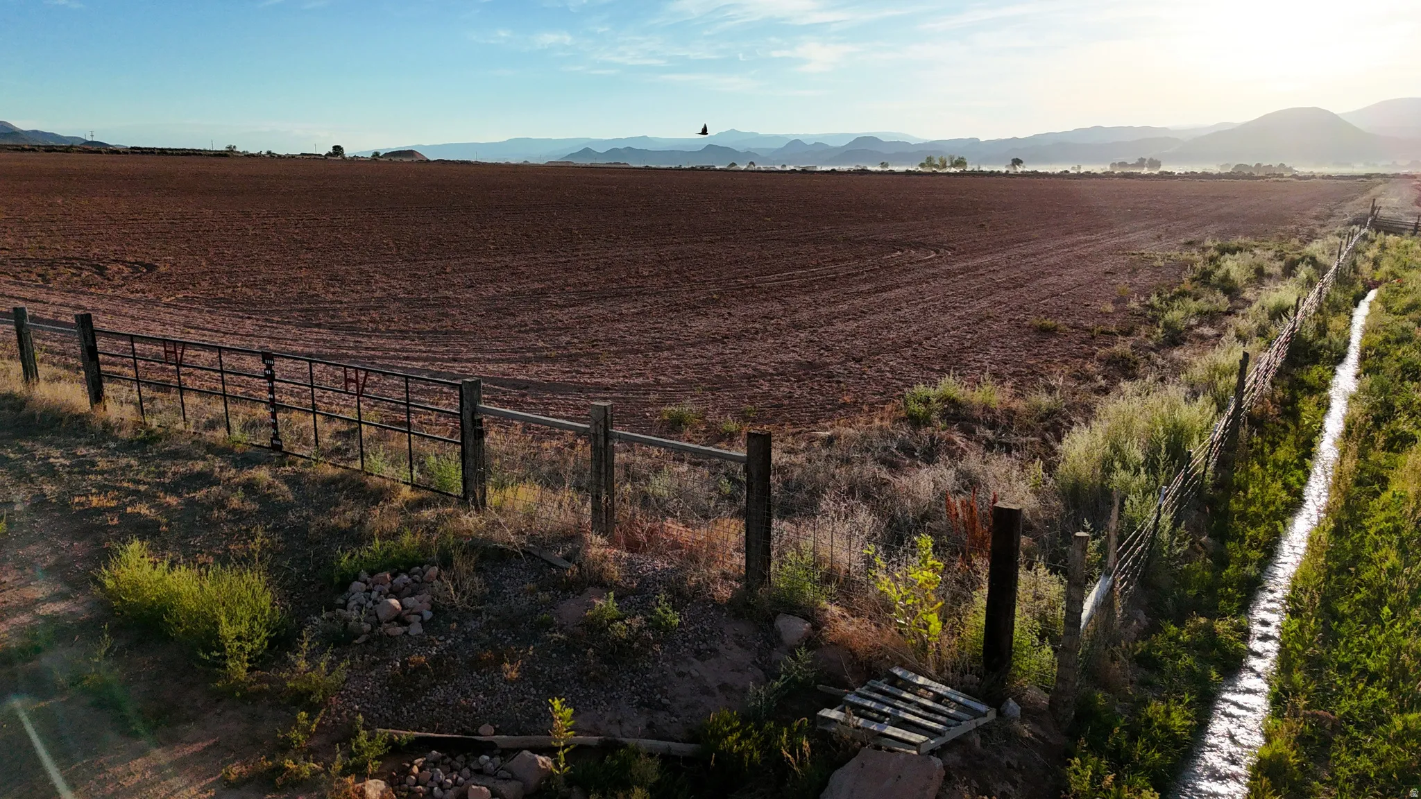 Expansive open land with tilled soil, perimeter fencing, and a visible irrigation ditch