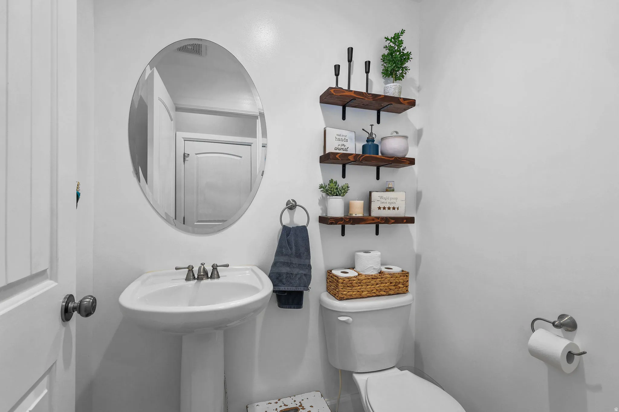 Pedestal sink with dual-handle faucet, oval mirror, wall-mounted shelving with dark wood finish, and a toilet with an adjacent paper holder