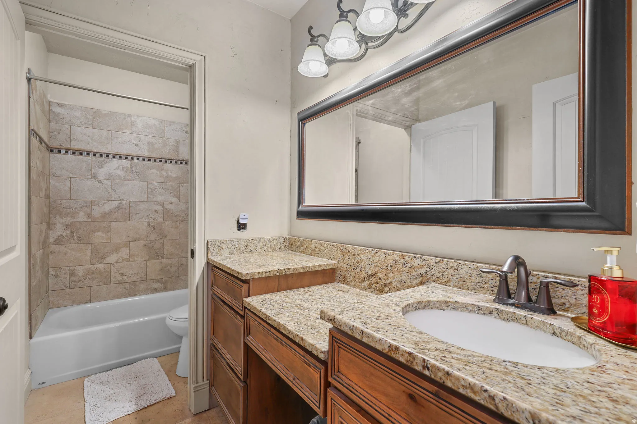 Wood-finish vanity with an integrated sink, stone countertop, oil-rubbed bronze faucet, large framed mirror, and tiled shower surround