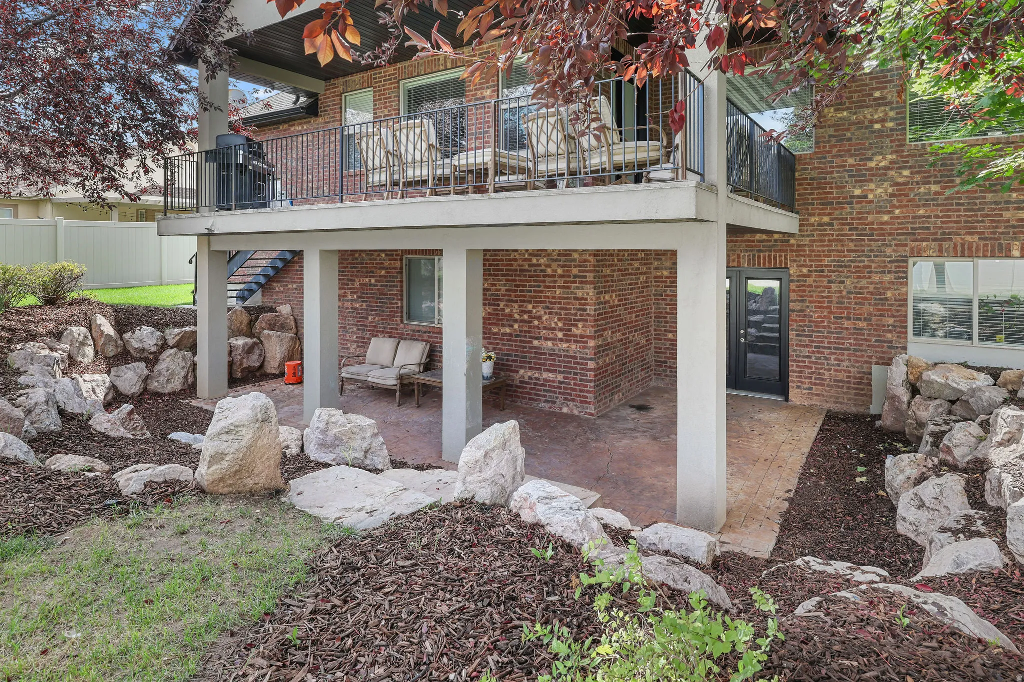 Two-story brick exterior with an upper-level balcony featuring a black metal railing, a covered patio below, and a landscaped yard with rock accents