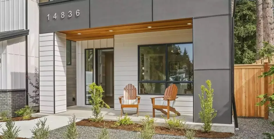 Modern exterior featuring horizontal white siding, dark gray paneling, natural wood soffit, black-framed windows, and a concrete pathway with gravel landscaping