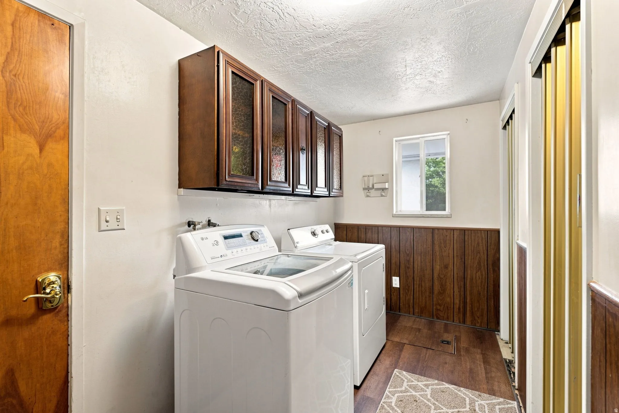 Laundry area featuring wood-finish flooring, half-wall wood paneling, and upper cabinetry with glass inserts
