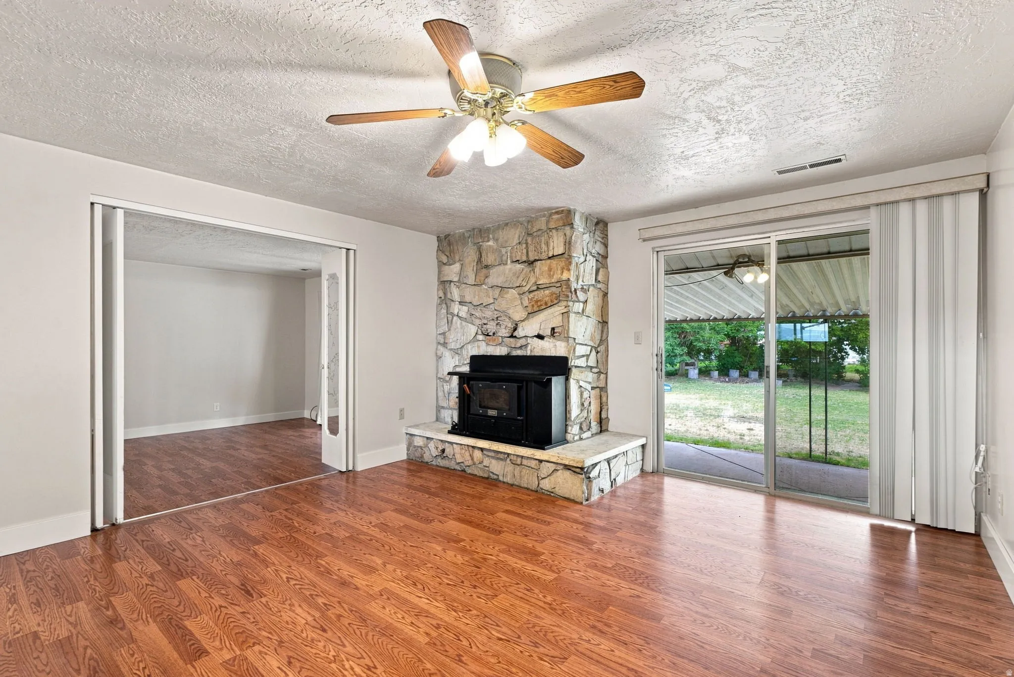 Wood-finish flooring, stone fireplace with a black wood-burning insert, ceiling fan, and sliding glass doors providing access to the yard