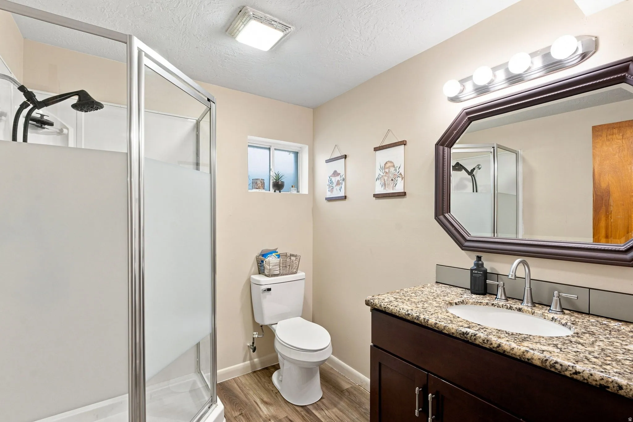 Wood-finish flooring with a glass-enclosed shower, single vanity with a granite-style countertop, and an octagonal framed mirror