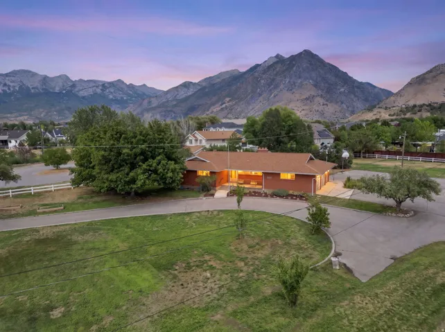 Brick facade residence with an attached garage, expansive paved driveway, surrounding green landscape, and distant mountain backdrop
