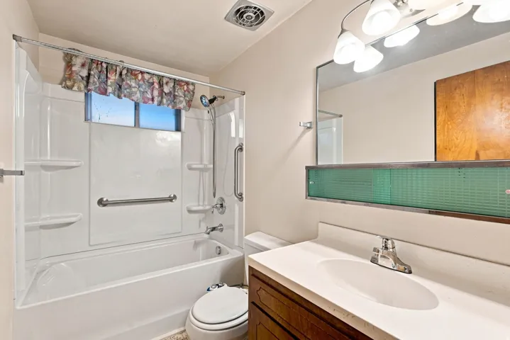 Full bath featuring a white tub and shower surround, wall-mounted grab bars, a built-in vanity with single basin sink, and an integrated mirror with overhead lighting