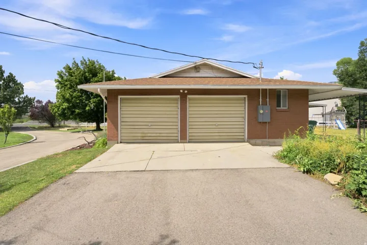 Brick exterior with dual-bay garage featuring paneled doors, concrete driveway, and mature landscaping with established trees and shrubs throughout the grounds