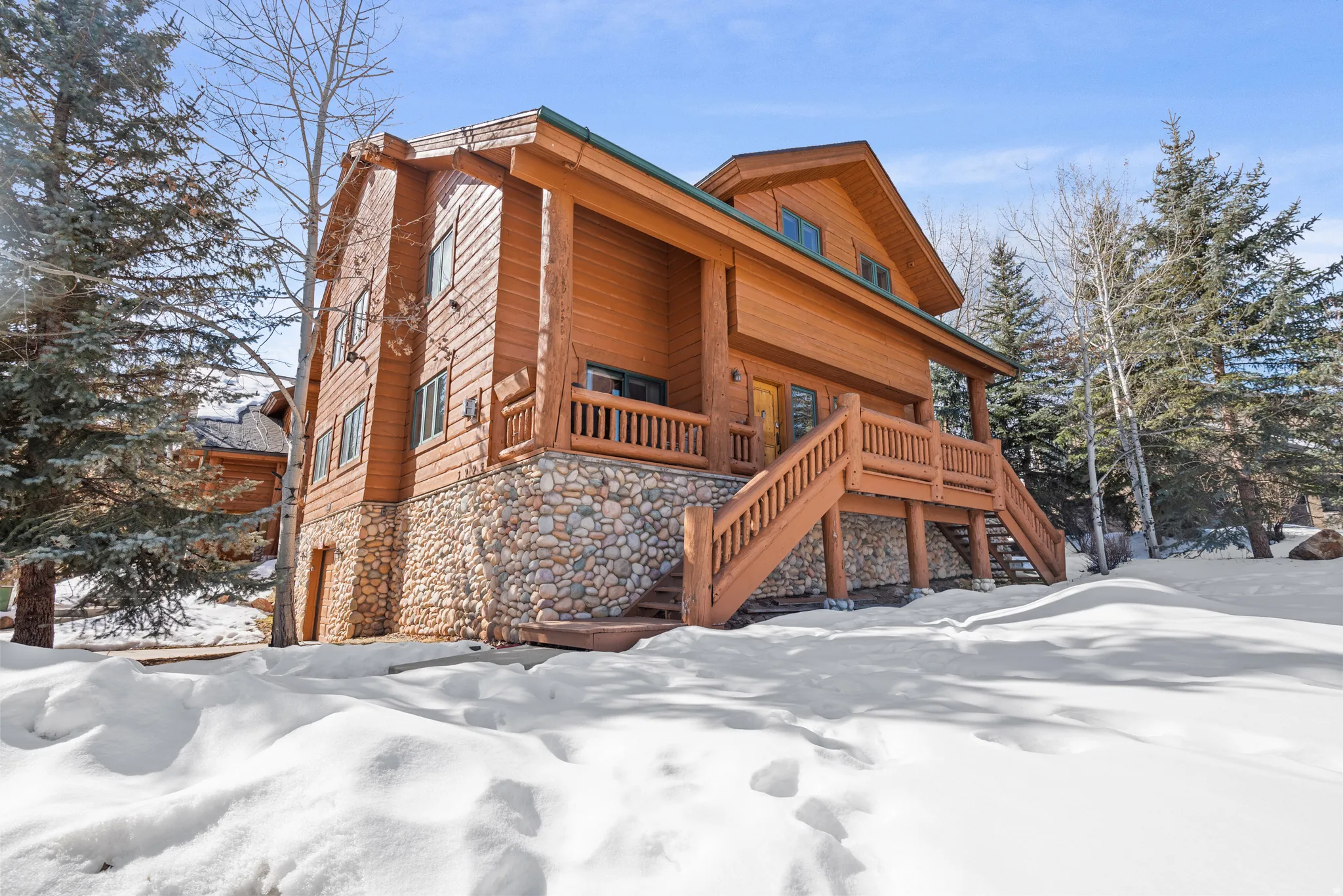 Log cabin exterior with natural wood siding, stone foundation, multi-story structure, expansive deck, and surrounding evergreen trees