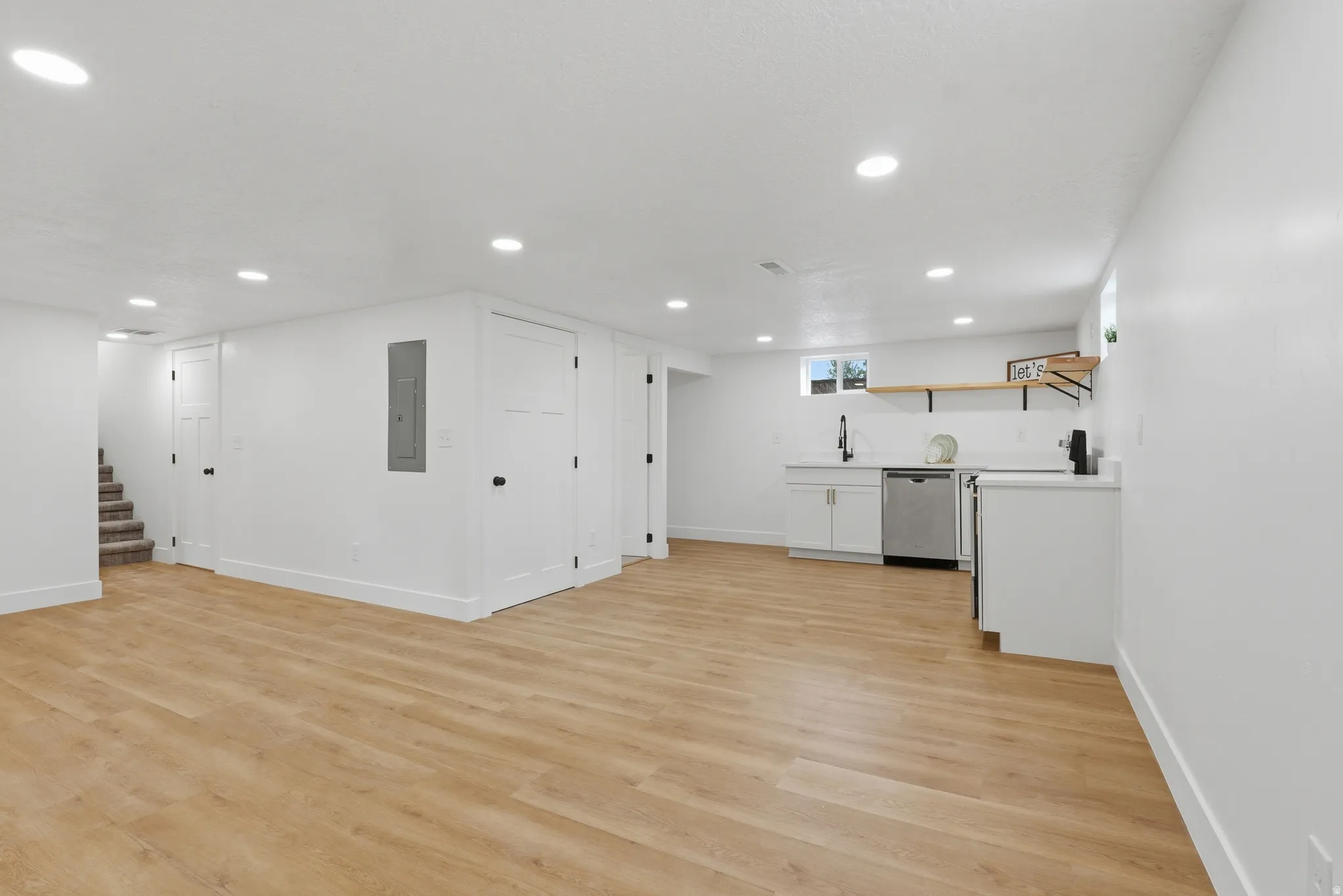 Expansive open room featuring wood-finish flooring, recessed lighting, a built-in wet bar with stainless steel appliances, and floating wood shelving