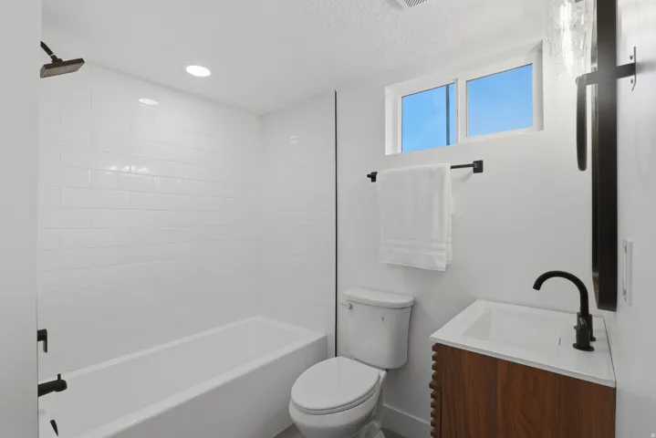 White subway tile shower surround with a dark bronze rainfall shower head, wood-finish vanity with a white countertop and a dark bronze faucet, and a horizontally oriented window