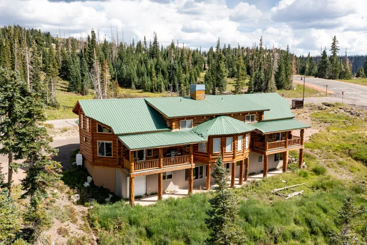 Log cabin exterior with a green metal roof, multiple balconies, and a surrounding natural landscape with mature evergreen trees