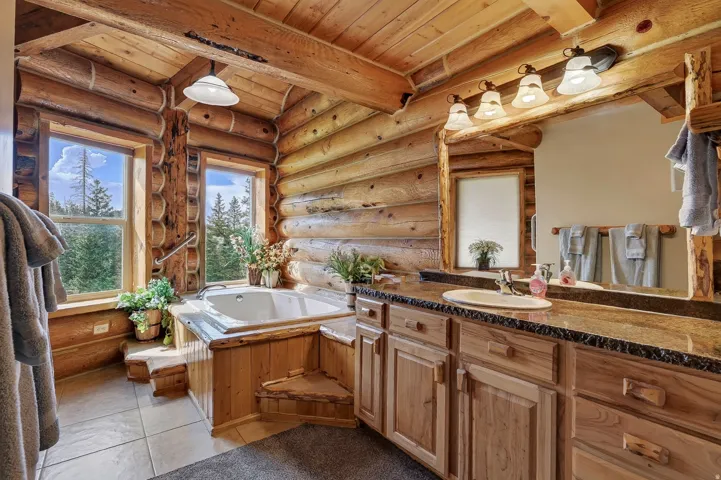 Log-clad interior featuring a built-in jetted tub, wood-finish flooring, a double vanity with a stone-finish countertop, and multiple windows