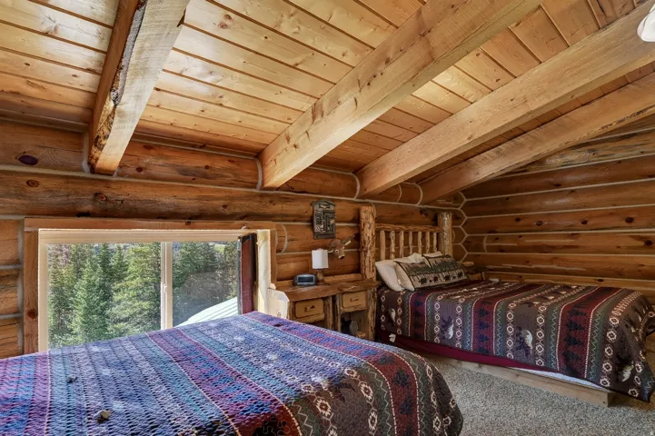 Rough-hewn log walls, exposed wood beam ceiling, wood-finish slatted ceiling, window with outdoor scenery, and light-colored carpet flooring