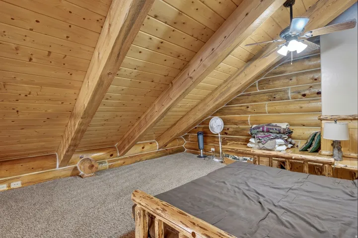 Vaulted wood-paneled ceiling, exposed wood beams, log walls, light carpet flooring, and a ceiling fan