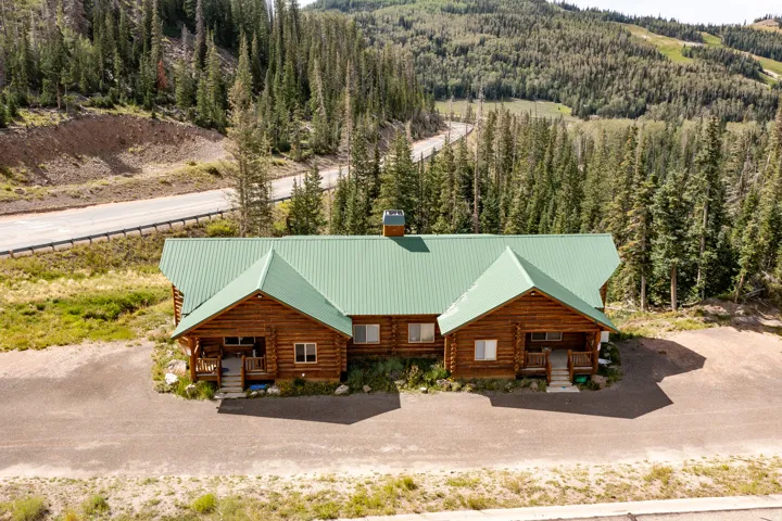 Log cabin exterior with a green metal roof, multiple covered entryways, and a surrounding forest landscape