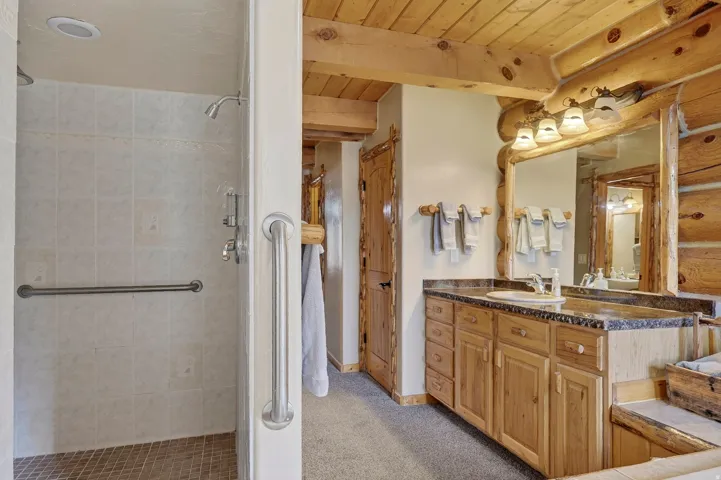 Wood-finish log wall construction, natural wood ceiling, wood-finish vanity with countertop, framed mirror with vanity lighting, and a tiled shower with grab bars