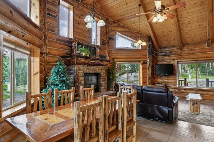 Vaulted wood-finish ceilings, prominent wood-finish beams, stone-clad fireplace with mantel, tiled flooring, and multiple windows