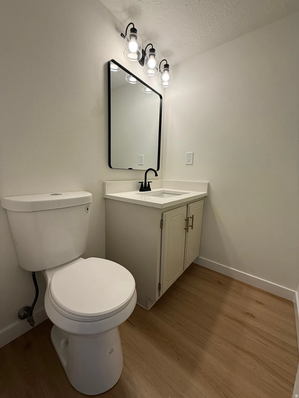 White vanity with countertop, framed mirror, black fixtures, and wood-finish flooring