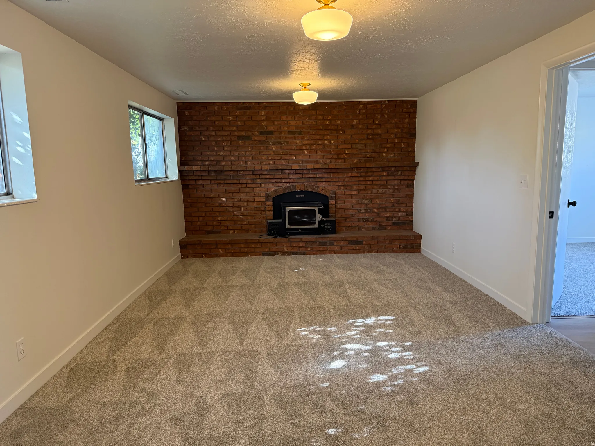 Brick accent wall with a built-in fireplace, two windows, two flush-mount ceiling lights, and patterned carpet flooring