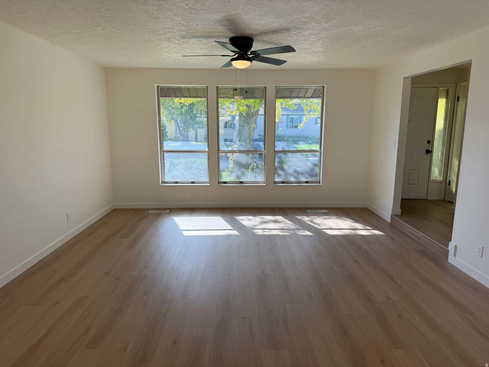 Spacious room with wood-finish flooring, three large windows, a ceiling fan, and light-toned walls