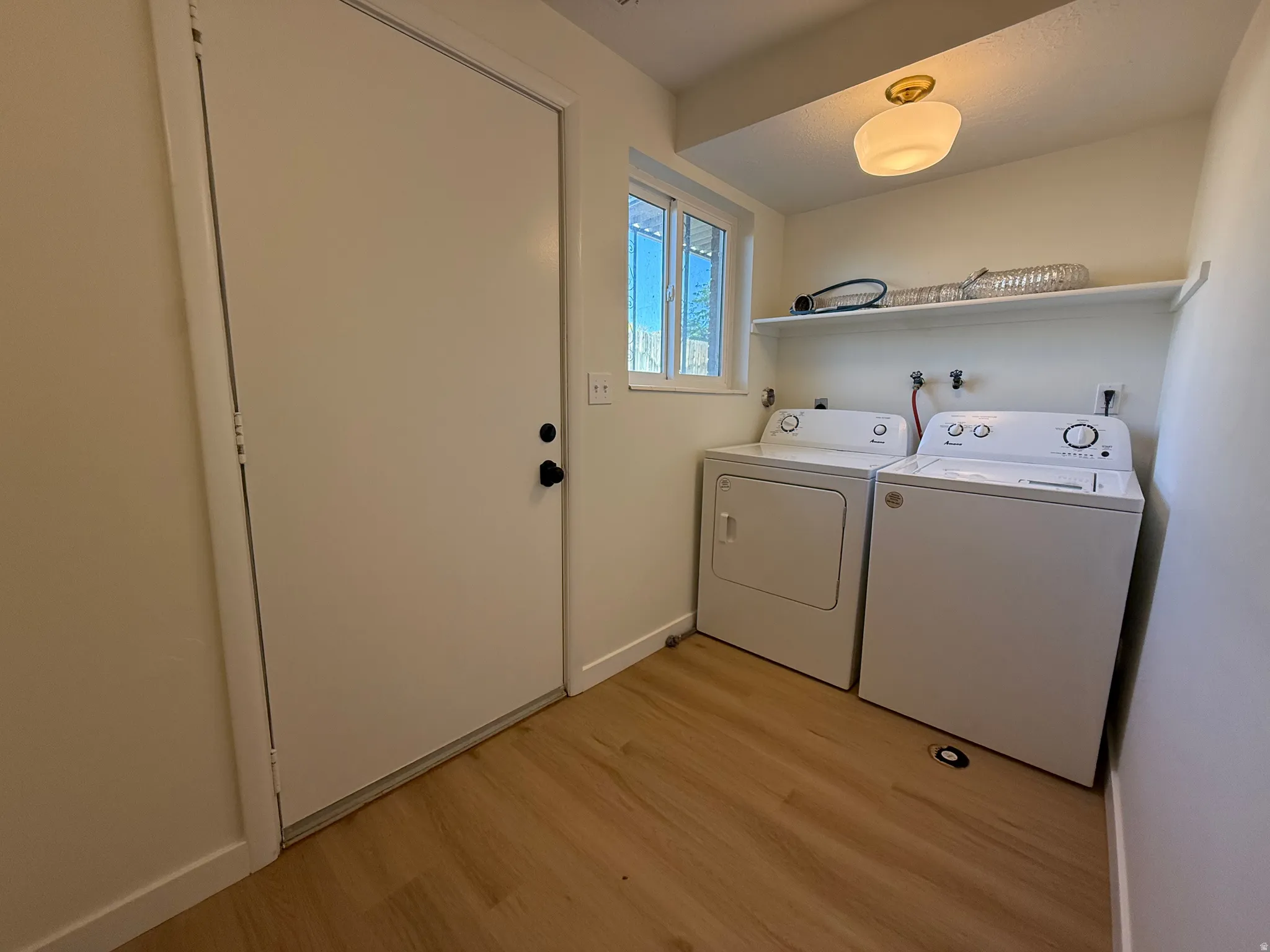 Laundry area with a white door, a window with horizontal panes, wood-finish flooring, a white wall-mounted shelf, and a ceiling-mounted light fixture