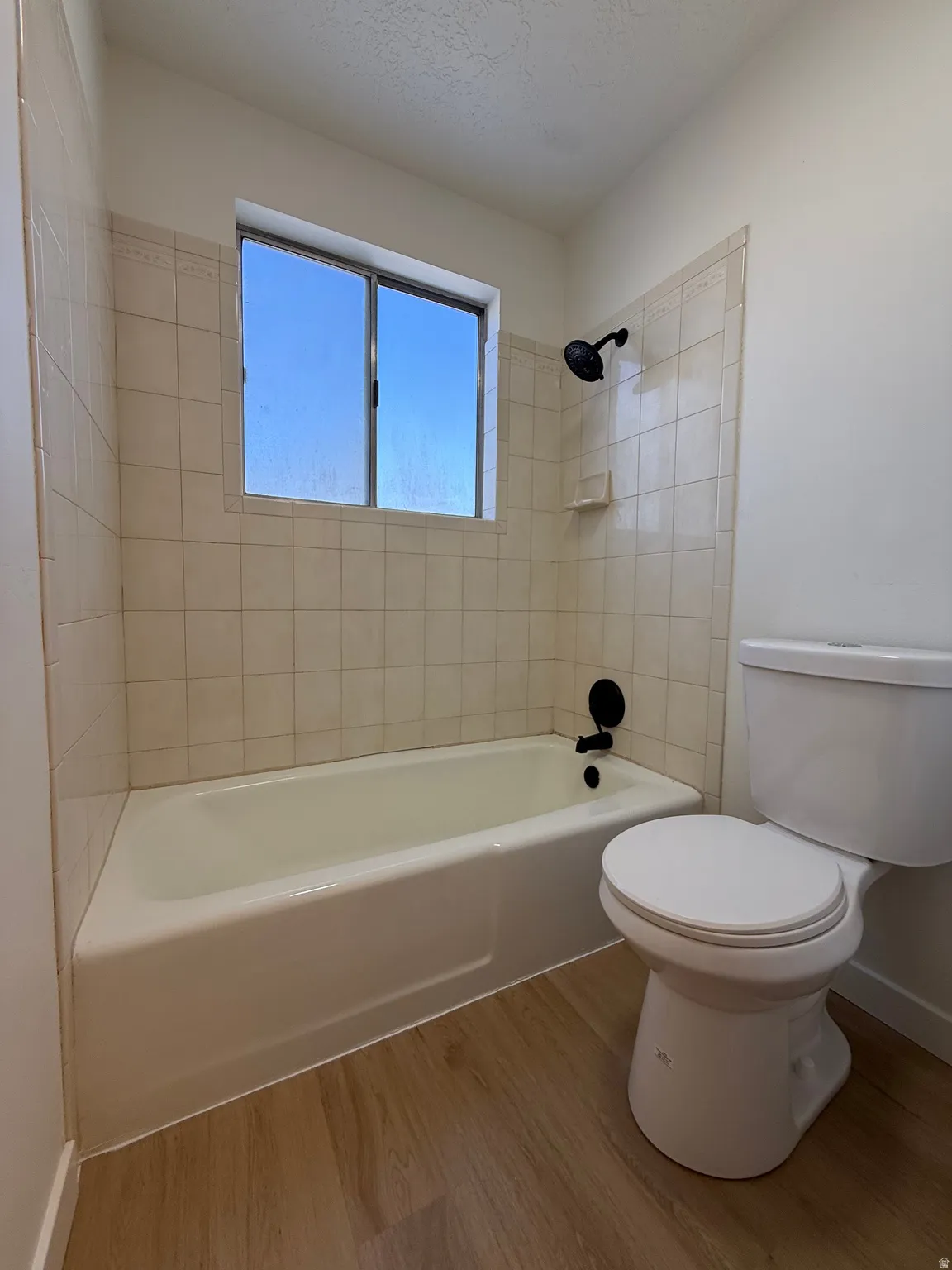 Bathtub with tiled surround, wood-finish flooring, a toilet, and a window