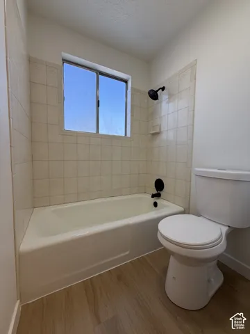 Bathtub with tiled surround, wood-finish flooring, a toilet, and a window
