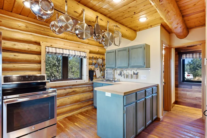 Rustic log cabin kitchen featuring exposed beam ceiling, stainless steel appliances, custom blue cabinetry with contrasting countertops, wood-finish flooring throughout, and pendant lighting suspended from timber beams