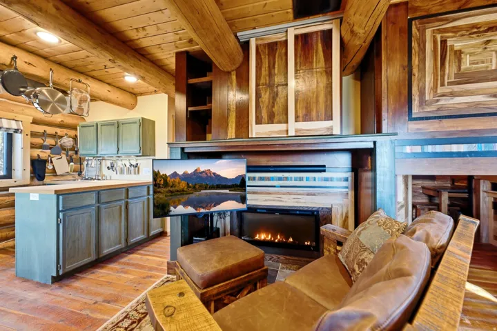Rustic interior featuring exposed log beams, wood-finish flooring, a built-in electric fireplace, and a kitchen island with light green cabinetry and a wood countertop