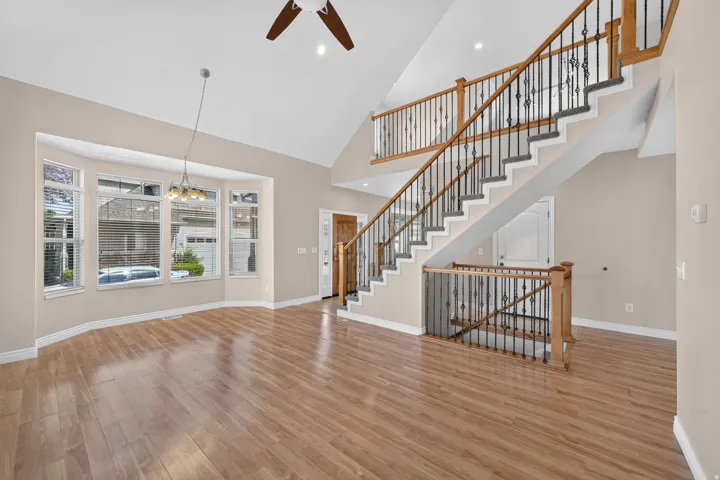 Vaulted ceilings, wood-finish flooring, bay window with blinds, dual-level staircase with decorative iron balusters, and a ceiling fan with integrated lighting