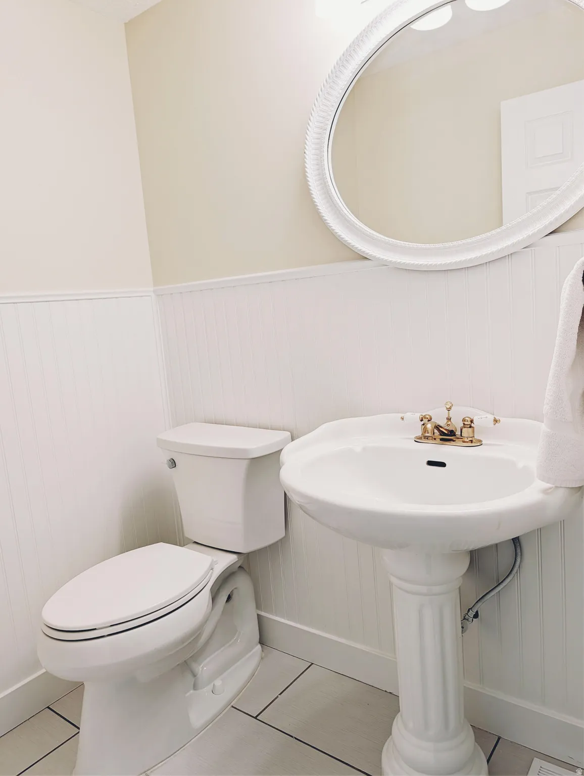 Pedestal sink with brass-finish faucet, elongated toilet, beadboard wainscoting, and large oval mirror