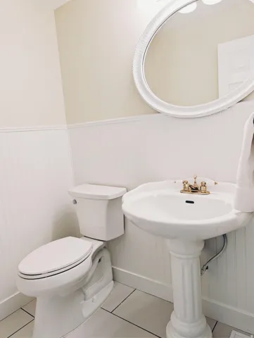 Pedestal sink with brass-finish faucet, elongated toilet, beadboard wainscoting, and large oval mirror