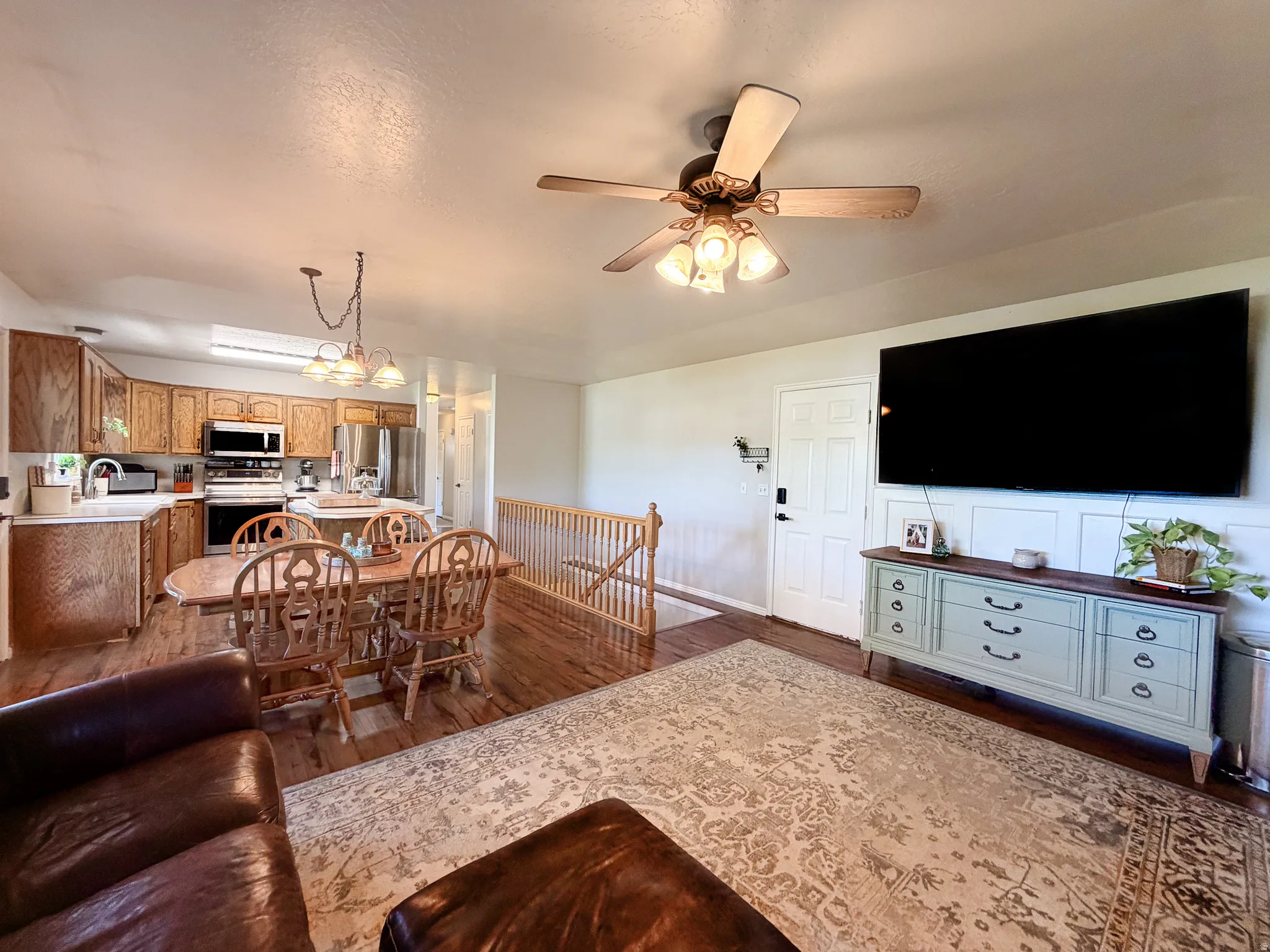 Wood-finish flooring, ceiling fan with integrated lighting, kitchen with wood cabinetry, stainless steel appliances, and a wall-mounted paneling accent