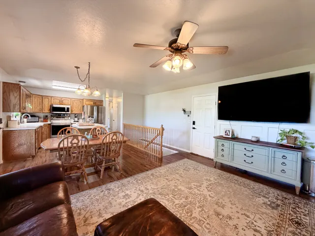 Wood-finish flooring, ceiling fan with integrated lighting, kitchen with wood cabinetry, stainless steel appliances, and a wall-mounted paneling accent